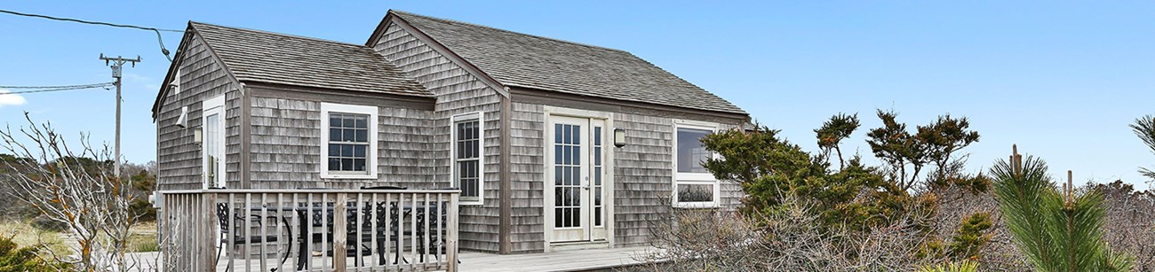 A wooden shingle house with a wide patio sits atop a sandy bluff.