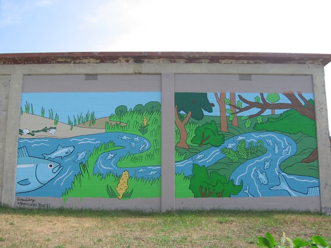Herring Revival Web A brick wall covered with a colorful mural depicting grey fish swimming up a river.