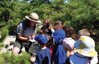 A female ranger in a sandy area with green low lying trees shows children something in a notebook.