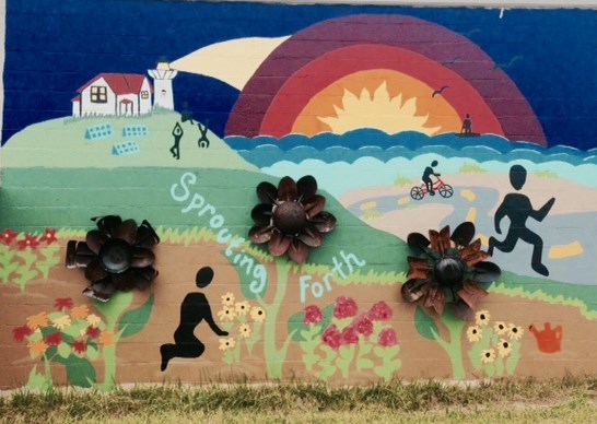 A brick wall with a colorful mural of black silhouettes engaged in a variety of activities on a hill by the ocean. There are four welded metal flowers as part of the mural.