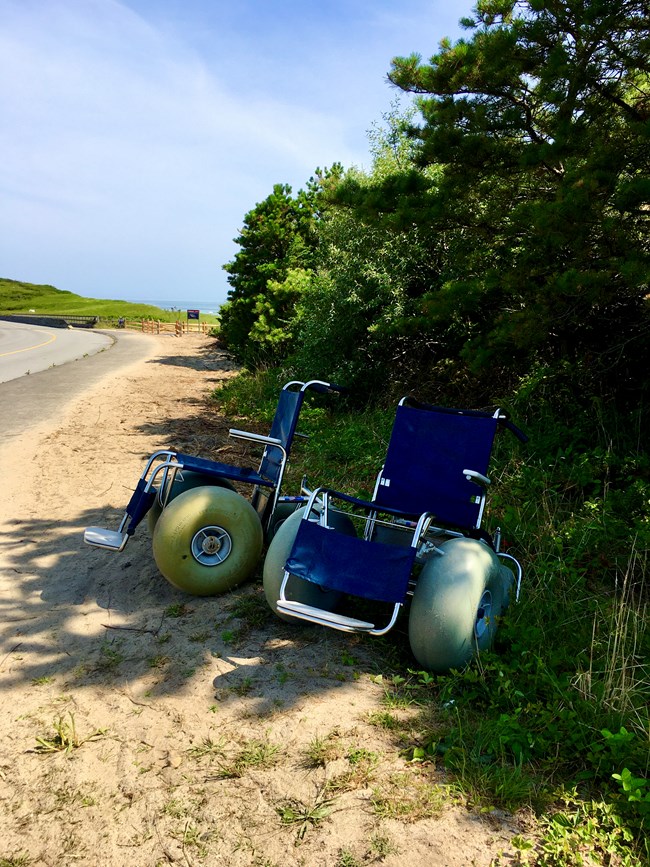 Two wheelchairs with large balloon tires are parked in the shade