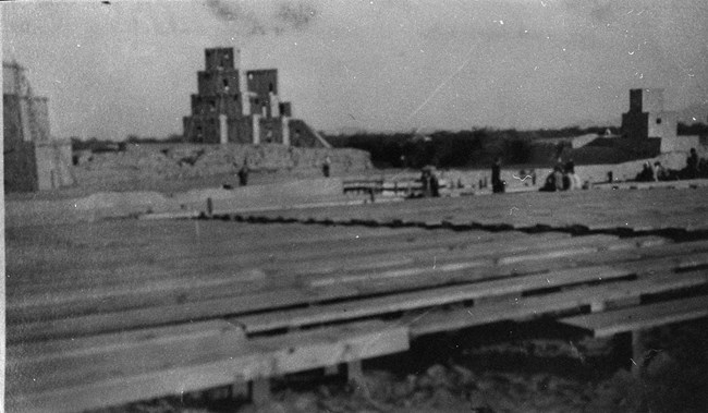 this black and white historic photograph shows the wooden plank seating facing a fake 'adobe' building on the earthern mound of compound b