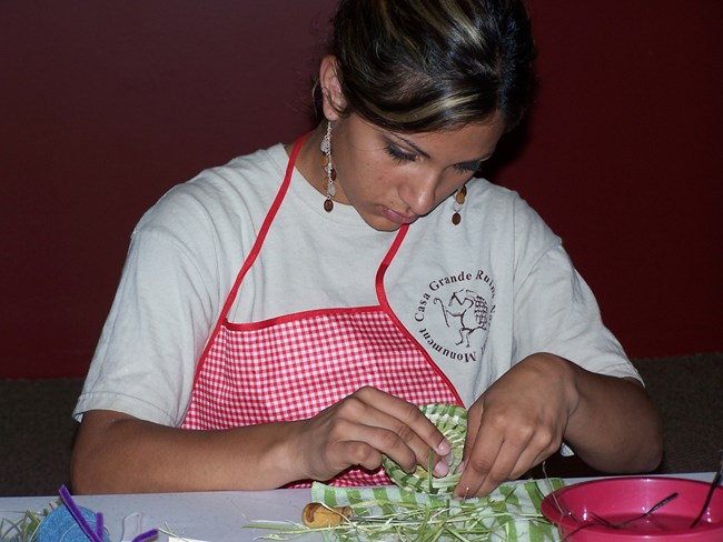 Sitting at a table with her work apron on YCC student works green yucca around the bear grass on the basket's edge