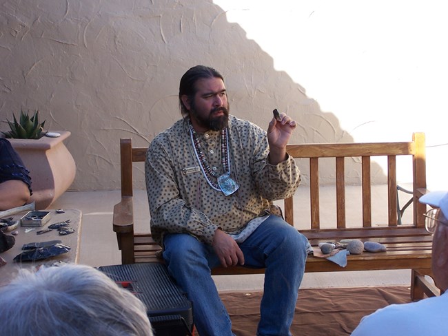 Ben sits on a shaded bench holding up a small projectile point he made from obsidian rock.