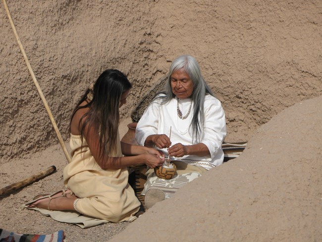 Costumed Actresses Mabelene and Cheynne sit in the corner of one of the smaller ruins spinning cotton fiber from a small basket onto a hand spindle