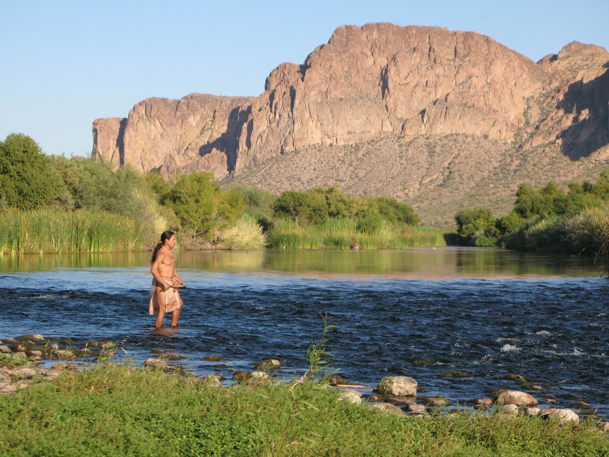 costumed actor Ruben Alvez stands in the Salt River with the Supersition Mountains in the background