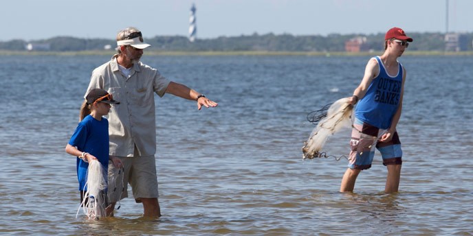 Volunteer and two visitors standing and cast-netting in the Pamlico Sound with the Cape Hatteras Lighthouse visible in the distance