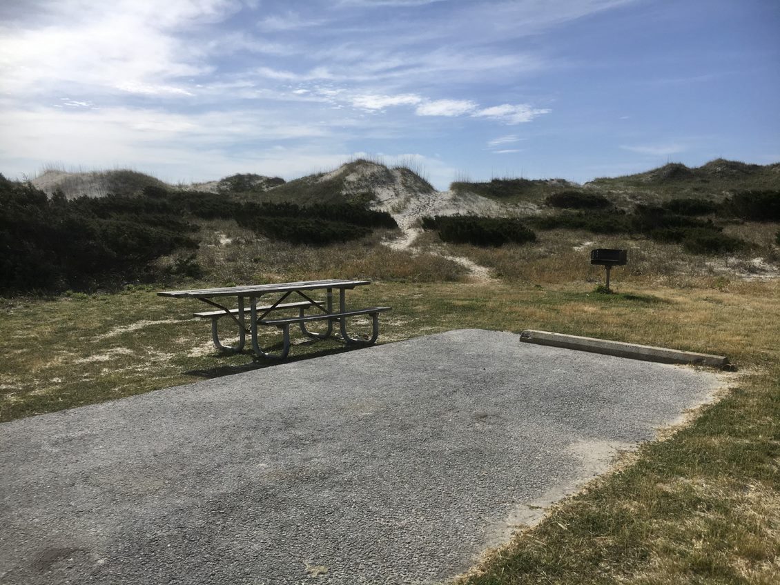 Standard camp site at Ocracoke Campground. Photo of camp site with picnic table, paved parking pad, charcoal grill, and dune in background.