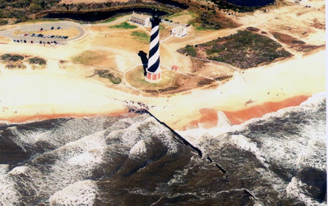 An aerial photograph shows the Cape Hatteras Lighthouse very close to the ocean waves.
