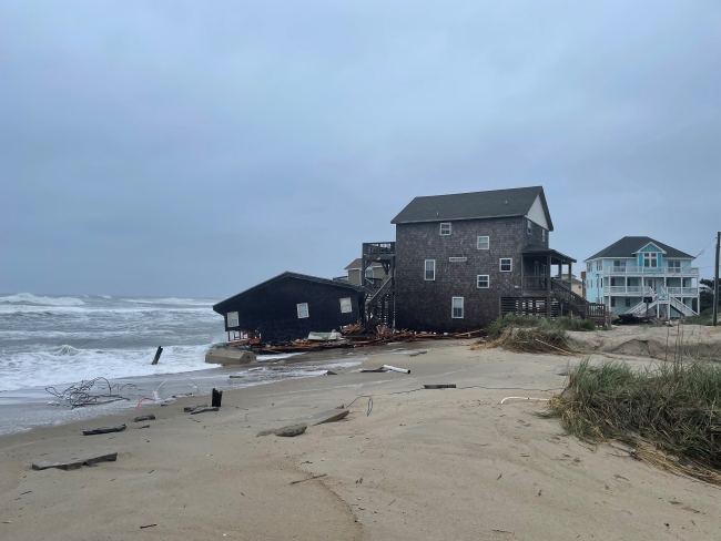 A portion of a collapsed house sits on beach next to ocean.