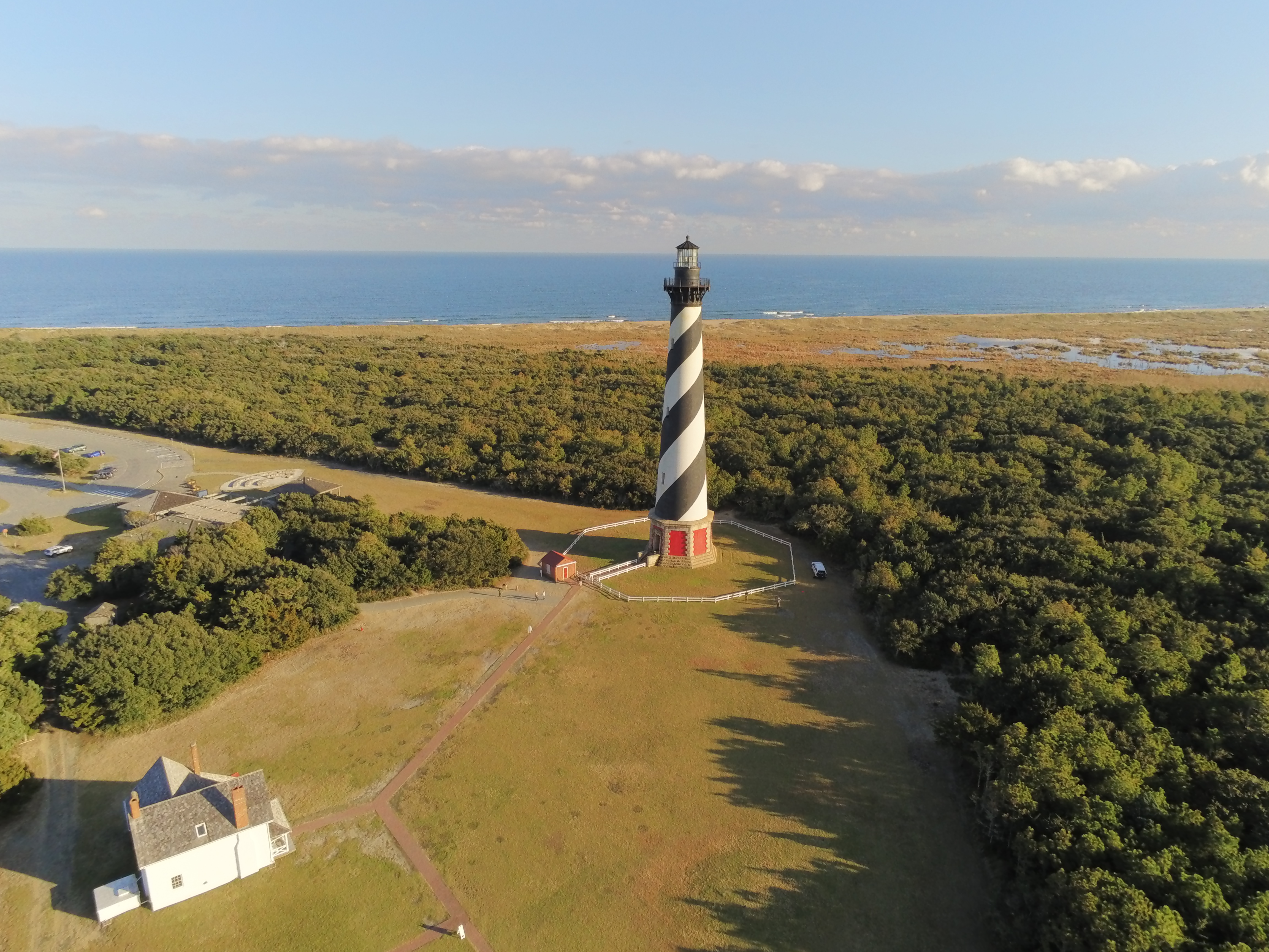 Aerial view of the Cape Hatteras Lighthouse, a white building, trees, and the ocean in background.