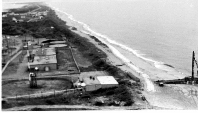 Black and white aerial photo showing white sandbags and construction equipment on the beach.