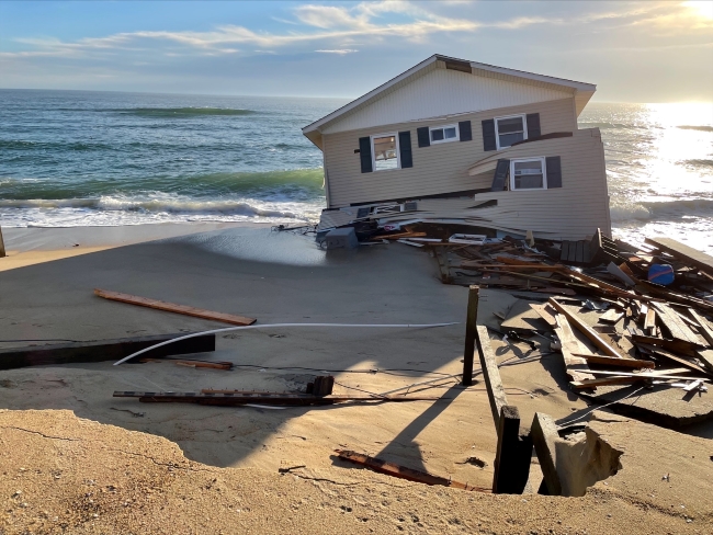 Collapsed house and debris on beach near the ocean.