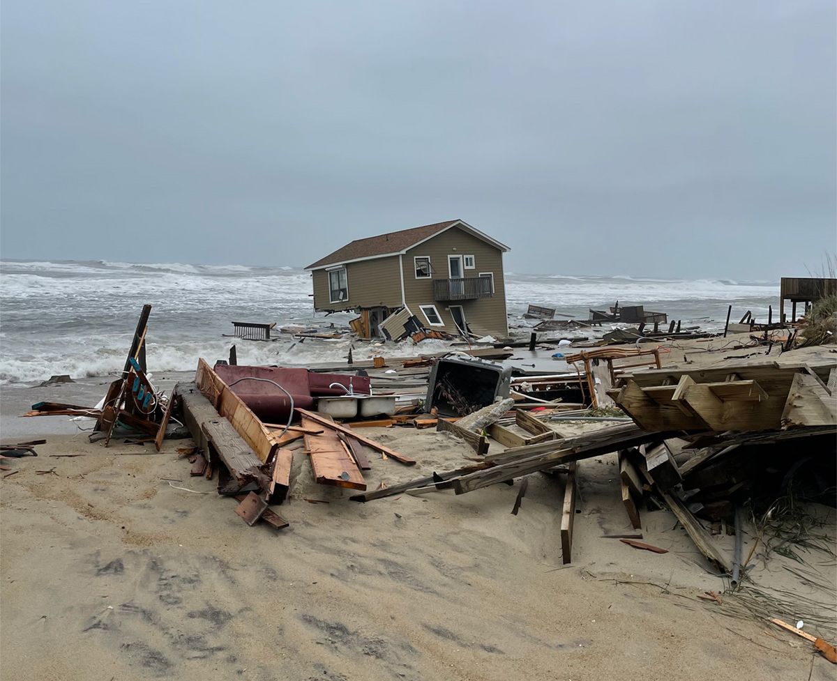 Collapsed oceanfront home. Home is seen submerged in water and the beach is scattered with debris.
