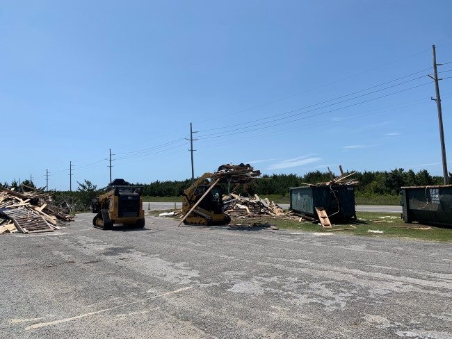 Two pieces of heavy equipment work to load beach debris into green dumpsters.