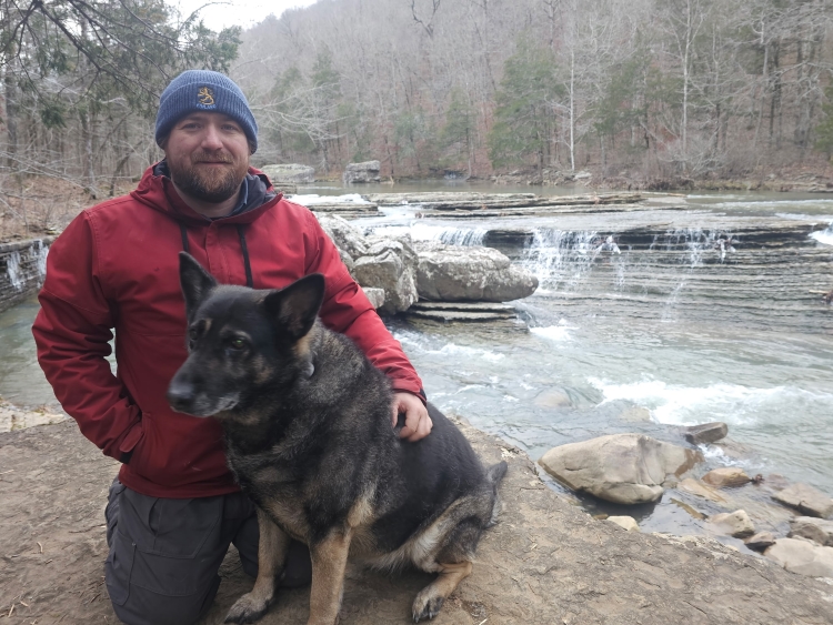 Photo of a bearded man with a blue winter hat, red jacket and kneeling next to a German shepherd dog.
