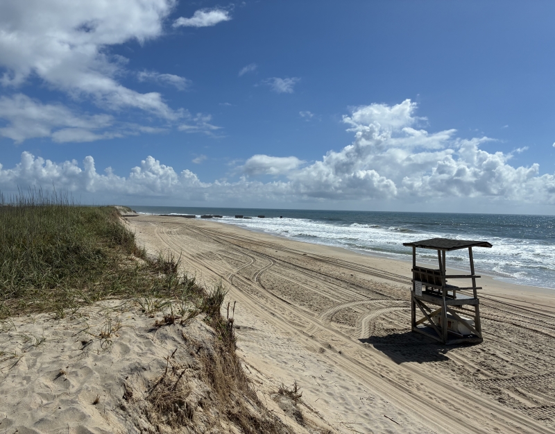 A wooden lifeguard stand with a sun shade stands in center of beach in front of ocean.