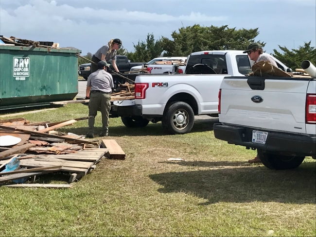 Three uniformed individuals work to offload collected wooden debris from white trucks.