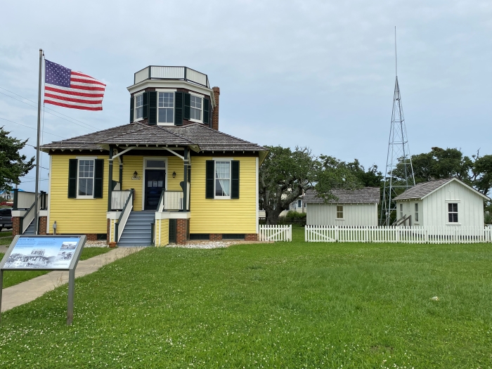 Photo of yellow building, U.S. flag, two white buildings, and a large metal tower.