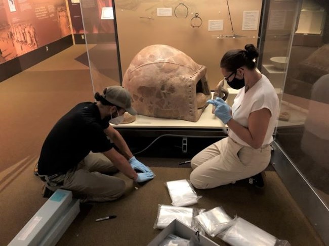 Two people kneel in front a museum exhibit and work on some artifacts.