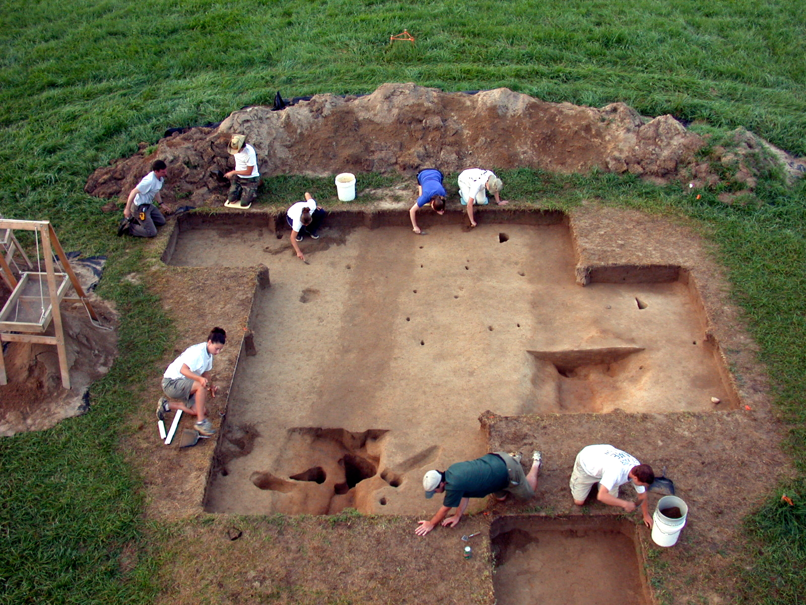 Eight archeologists carefully excavate a large pit at Werowocomoco that reveals dark stains in the earth. The stains indicate the presence of parallel trenches constructed in the Native site.