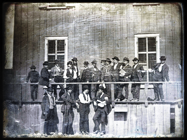 Historic image of men in suits, standing in front of a wooden building.