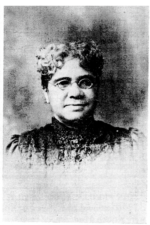 Faded image of the head and shoulders of a slightly smiling woman wearing wire-rimmed glasses and a high-necked, ruffled dress. Her hair is parted in the middle and pulled back, with loose curls on top of her head. Her face is turned slightly to her left.