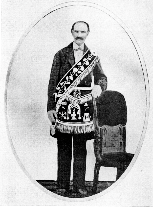 Man standing with his right arm at his side and left arm resting on the back of a chair. The man is light-skinned, balding, and has a thick mustache. He wears a dark suit with a white shirt and an elaborate embroidered bandoleer and apron with the symbols