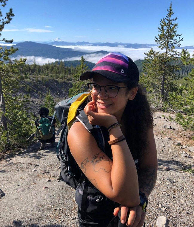 lt= A woman stands in front of a scenic forest and mountains.