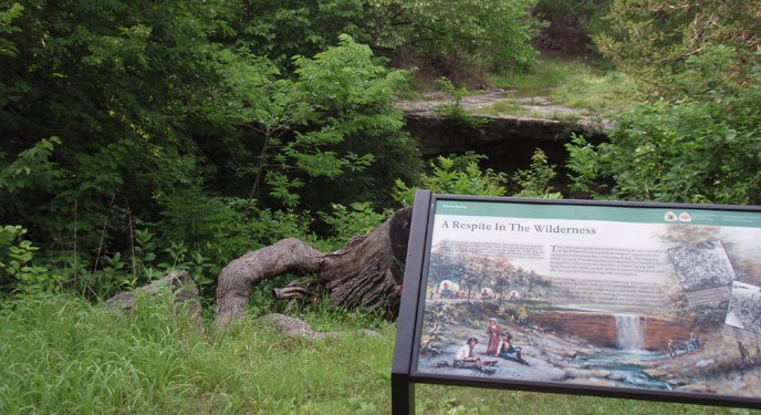 A wayside exhibit in front of Alcove Spring tells the story of emigrants who passed through this site in the 1800s. Wayside in front of green shrubs and grass, and a limestone ledge. A wayside exhibit in front of Alcove Spring tells the story of emigrants who passed through this site in the 1800s. Wayside in front of green shrubs and grass, and a limestone ledge.