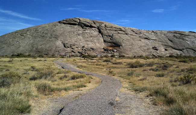 A hardened pathway leads through sagebrush to a large rounded grey rock formation with people in the distance. A hardened pathway leads through sagebrush to a large rounded grey rock formation with people in the distance.