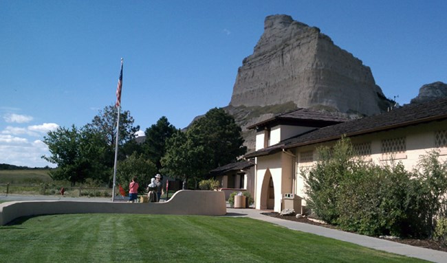 visitors stand in front of a cream building with brown shingles with a green lawn and a large rock buttress behind.