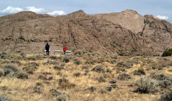 Two people stand in front of an outdoor exhibit while looking at a large rock formation called Split Rock in Wyoming with sagebrush. Two people stand in front of an outdoor exhibit while looking at a large rock formation called Split Rock in Wyoming with sagebrush.