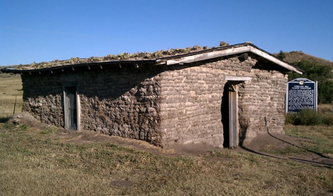 An old building made of sod bricks in a tan brown color with a roof covered in cactie sits on a grassy hill with blue sky above in Nebraska. An old building made of sod bricks in a tan brown color with a roof covered in cactie sits on a grassy hill with blue sky above in Nebraska.