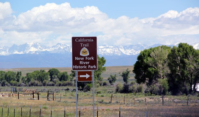 A brown directional highway sign with a yellow and white logo sits in front of cottonwood trees, and large snow covered gray mountains with clouds and blue sky
