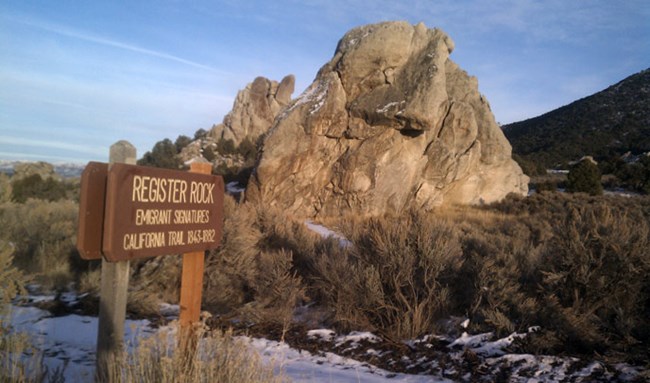 A large rock formation stands above sagebrush with a wooden engraved sign in front reading: "Register Rock Emigrant Signatures California Trail 1843-1882." A large rock formation stands above sagebrush with a wooden engraved sign in front reading: "Register Rock Emigrant Signatures California Trail 1843-1882."