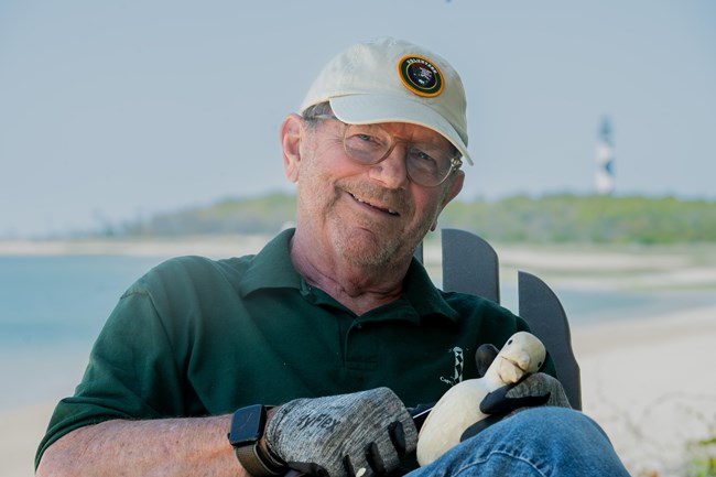 A man wearing glasses holds a shorebird decoy while sitting in a chair.