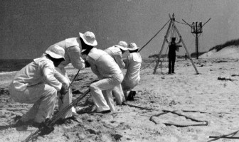 Black and white image of Beach Apparatus Drill reenactment. A group of surfmen are in the foreground pulling a rope on the sand. Another person stands in the background.