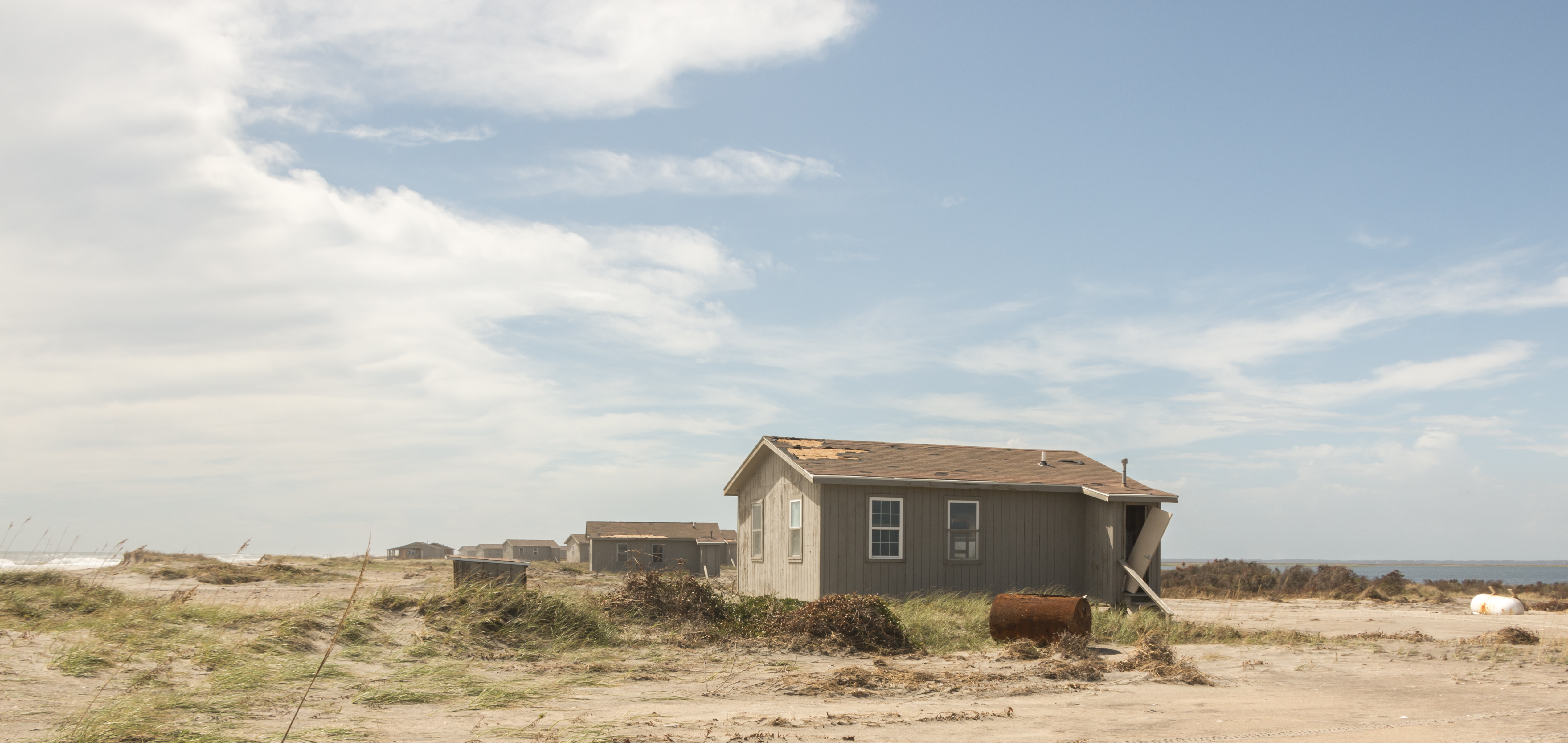 Looking down the beach at the edge of the dunes at a line of wooden cabins. The cabin in the forefront missing roof shingles and outside wood.