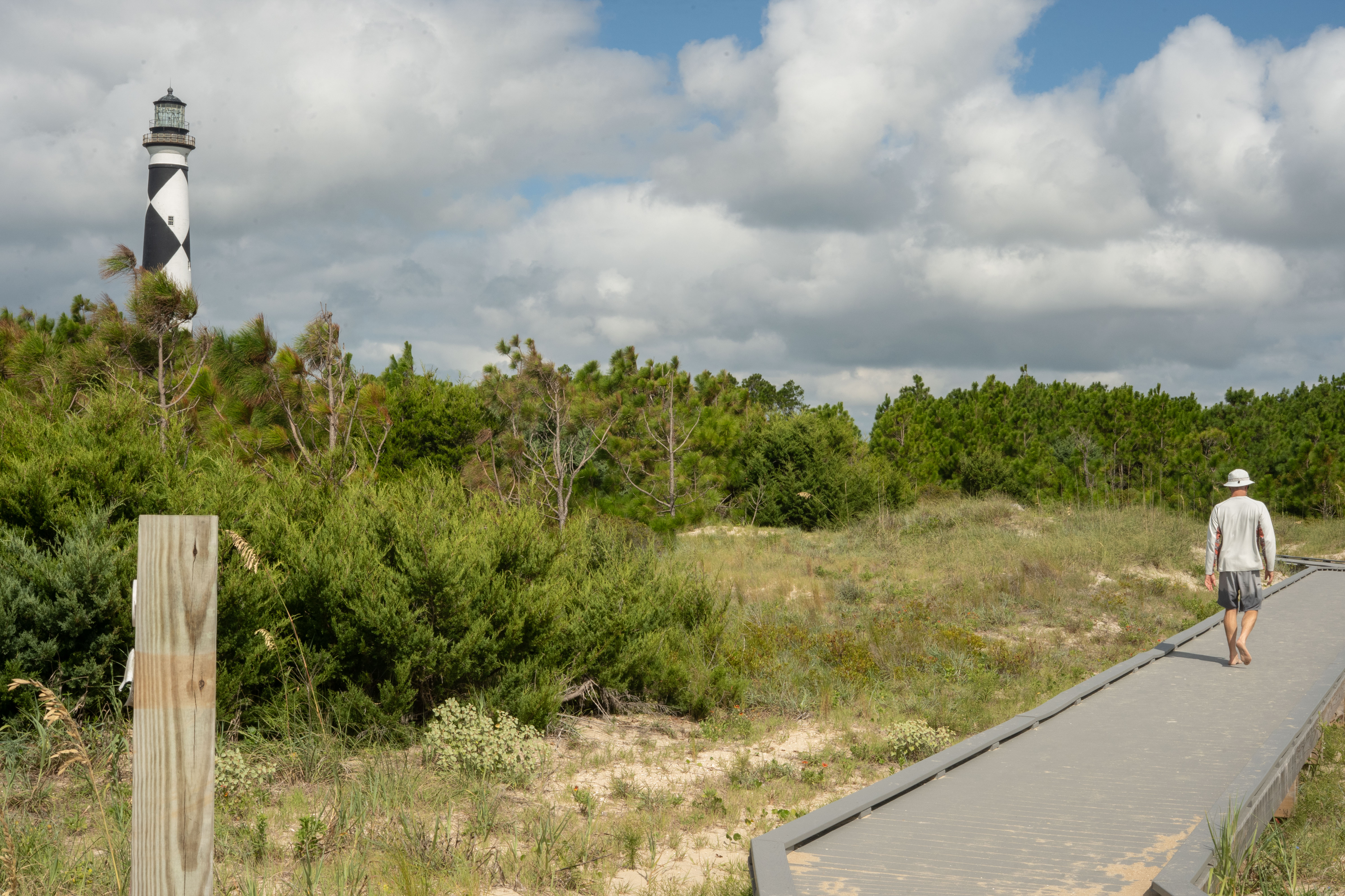 Hurricane_Erin20250822_SG_0146 A person walks on the boardwalk surrounded by trees. Lighthouse in the background.