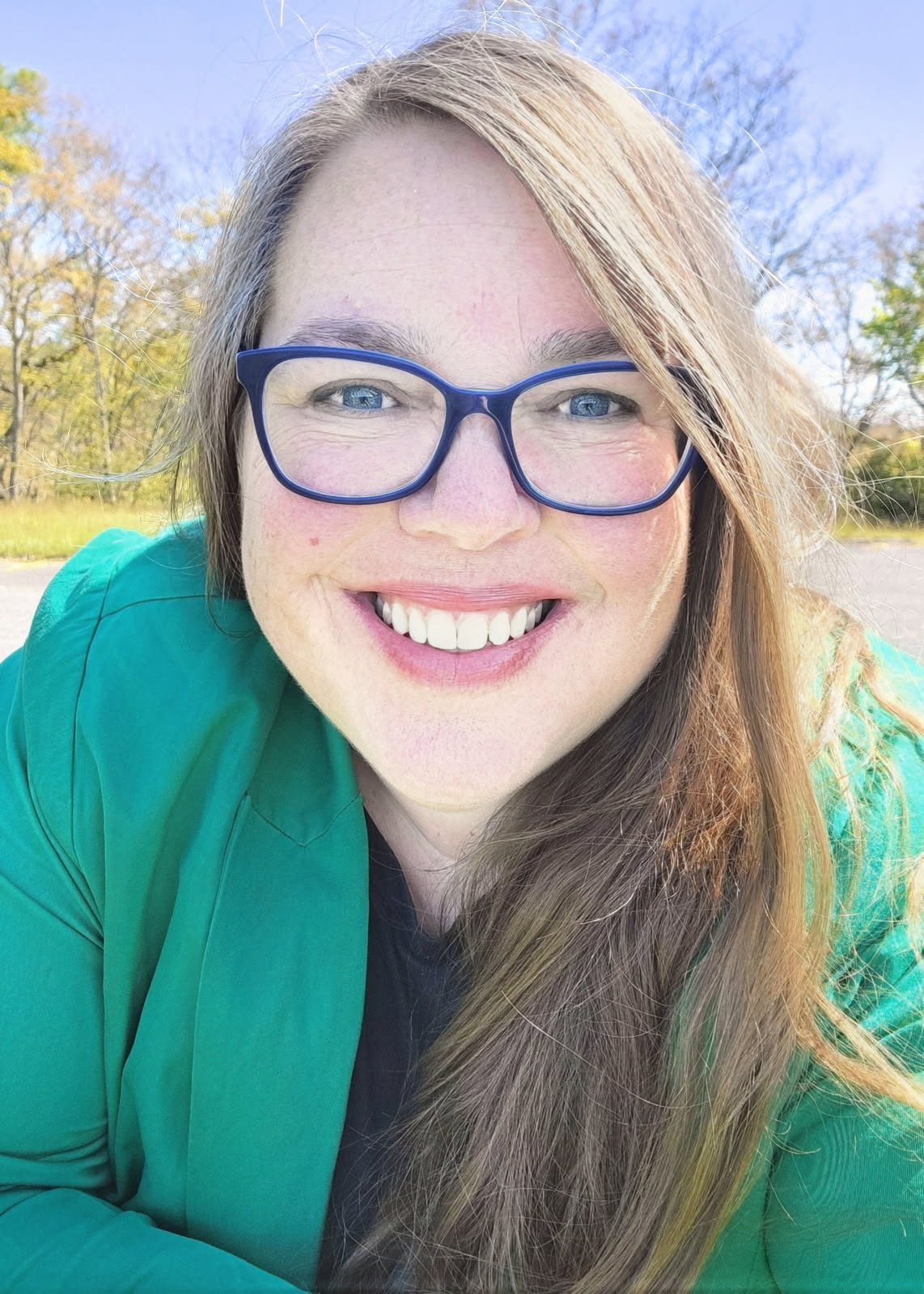 A woman wearing a green shirt and blue jacket takes a selfie.