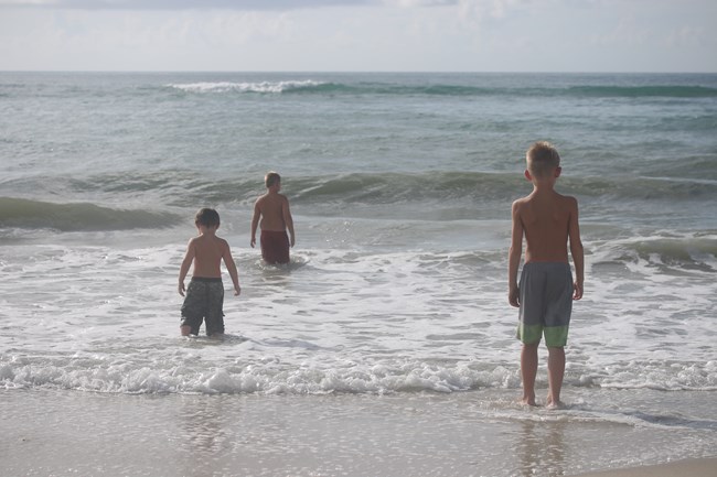 3 boys stand facing the water, wish waves craching towards them. Sand in the foreground.