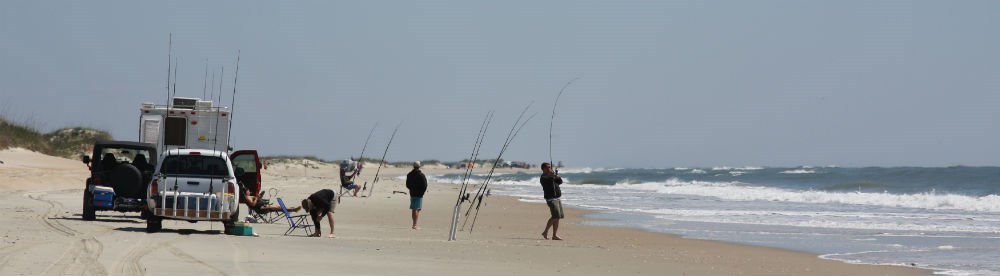 Two vehicles and a camper on the sand. Three visitors stand in between the vehicles and the ocean. Multiple fishing poles stand in the sand