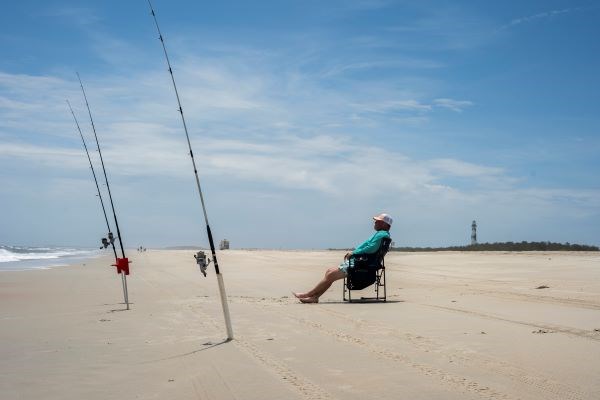 A person in a blue shirt, shorts, and a hat sits in a chair facing 3 fishing poles in the sand. A Vehicle in the background. Cape Lookout Lighthouse and pine trees in the back right.