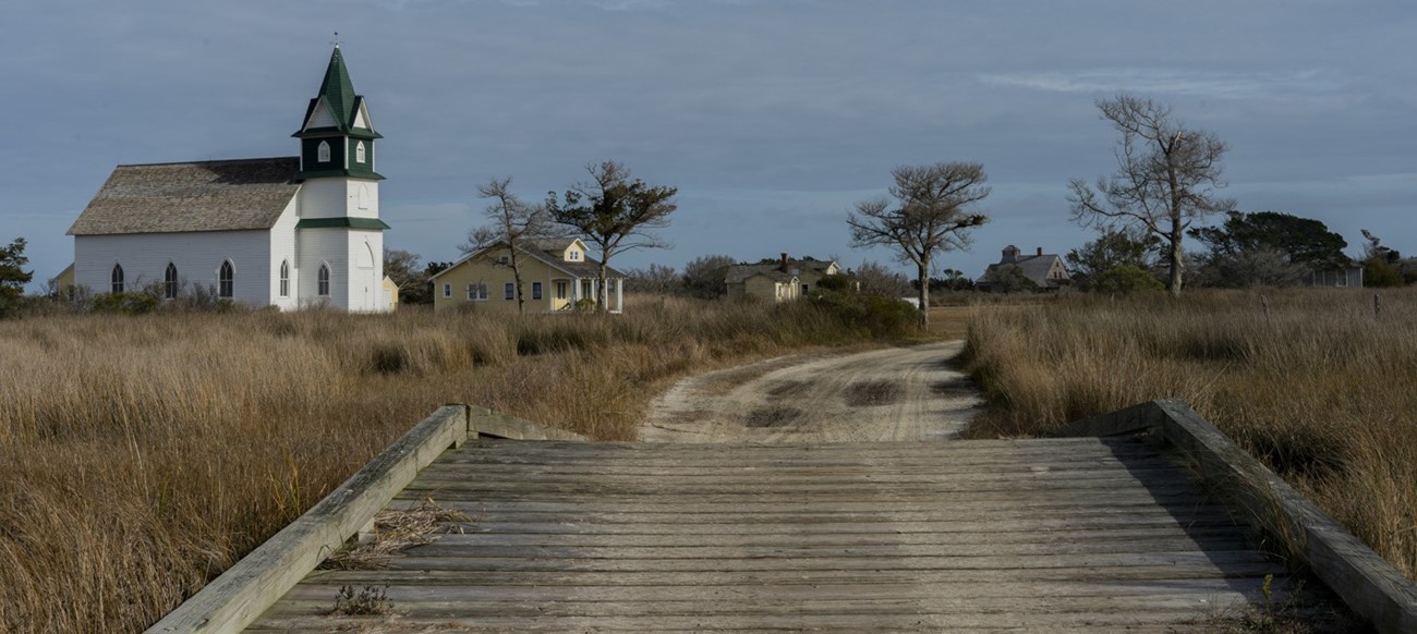 Wooden bridge leading to a church and houses on the right, and houses on the left. Trees and blue cloudy sky in the distance.