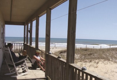 Visitors sit on wooden porch, with a sand dune to the right of them, and a blue sky behind them