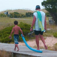 A man and boy walk along a boardwalk carrying beach toys.