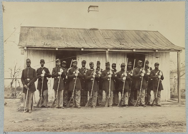 African American soldiers stand in formation in front of a wooden building during the Civil War.