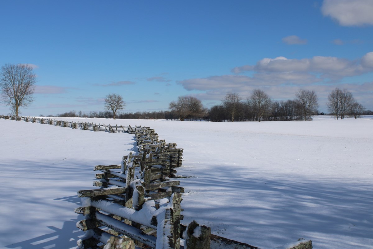 A wooden fence on a snow-covered field with leafless trees in the background.