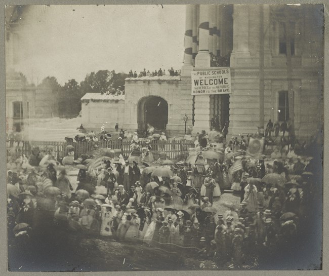 A crowd of people standing near a building with banner hung up on pillar.